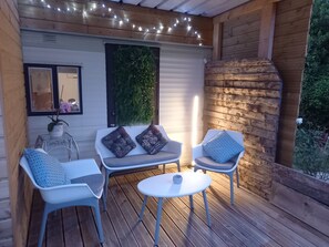A wooden deck with a seating area featuring two chairs and a small table, a decorative wall with greenery, and string lights. - Bungalow on a horse farm (Yèvres)