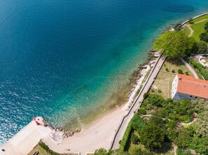 An aerial view of a coastal area with a clear beach, a paved walkway, and a building with a red roof. - Impeccable 4-bed Apartment in Osor (Mali Losinj)
