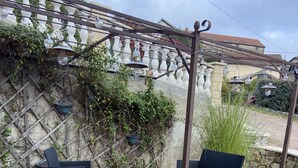 Een terras buiten met rieten meubilair, een pergola met hangende lichtjes en een stenen muur begroeid met klimplanten.