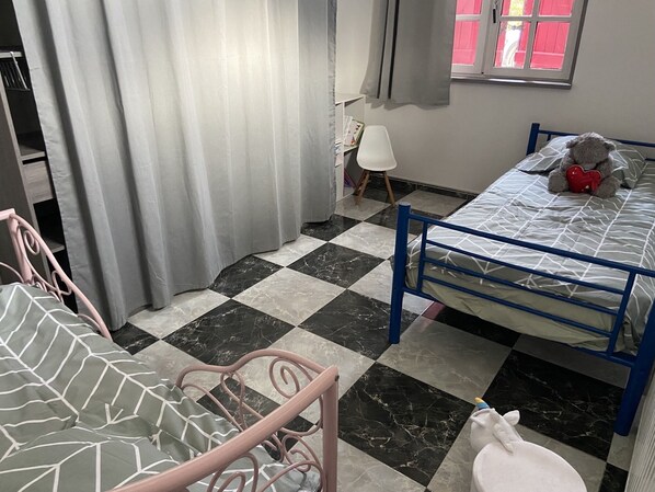 Une pièce avec un sol à carreaux, un lit avec un cadre en métal bleu, une chaise, une fenêtre avec des volets rouges et un rideau.