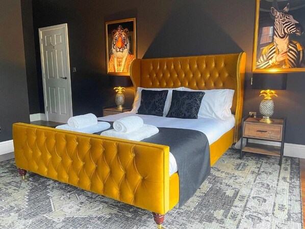 Una habitación con una cama de cabecera tapizada en amarillo, ropa blanca y dos toallas sobre la cama. Hay dos mesitas de noche con lámparas y una puerta al fondo.