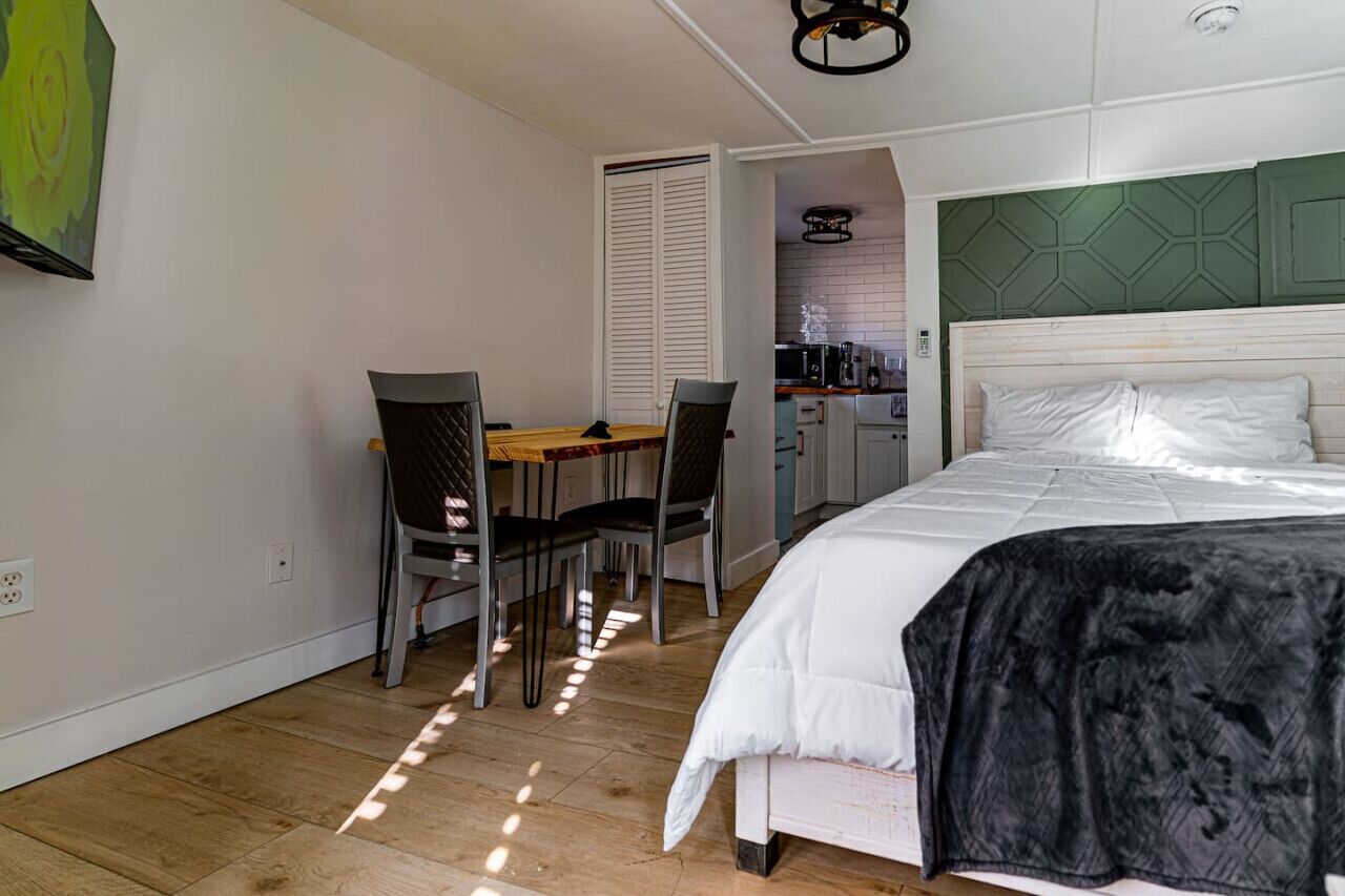 A bedroom with a bed, a dining table with chairs, and a kitchenette in the background.