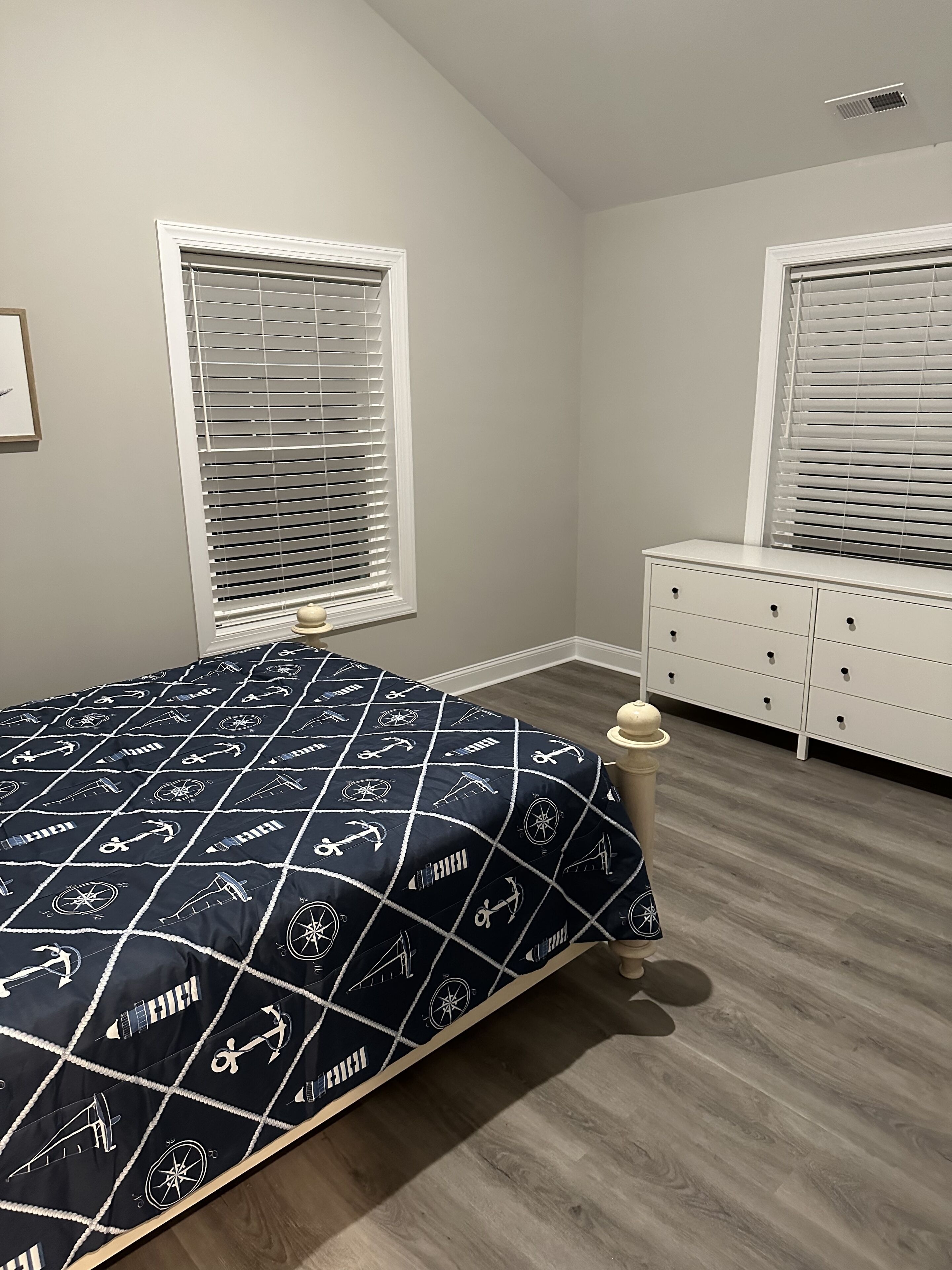 A bedroom with a bed, a dresser, two windows with blinds, and a ceiling vent.