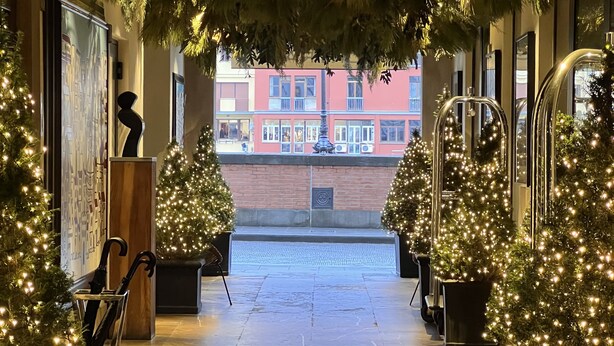 An indoor corridor with Christmas trees and lights, leading to a red building with balconies. - Oro Mono (Florence)