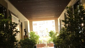 A corridor with a patterned ceiling, potted plants, and a view of a building in the distance. - Oro Mono (Florence)