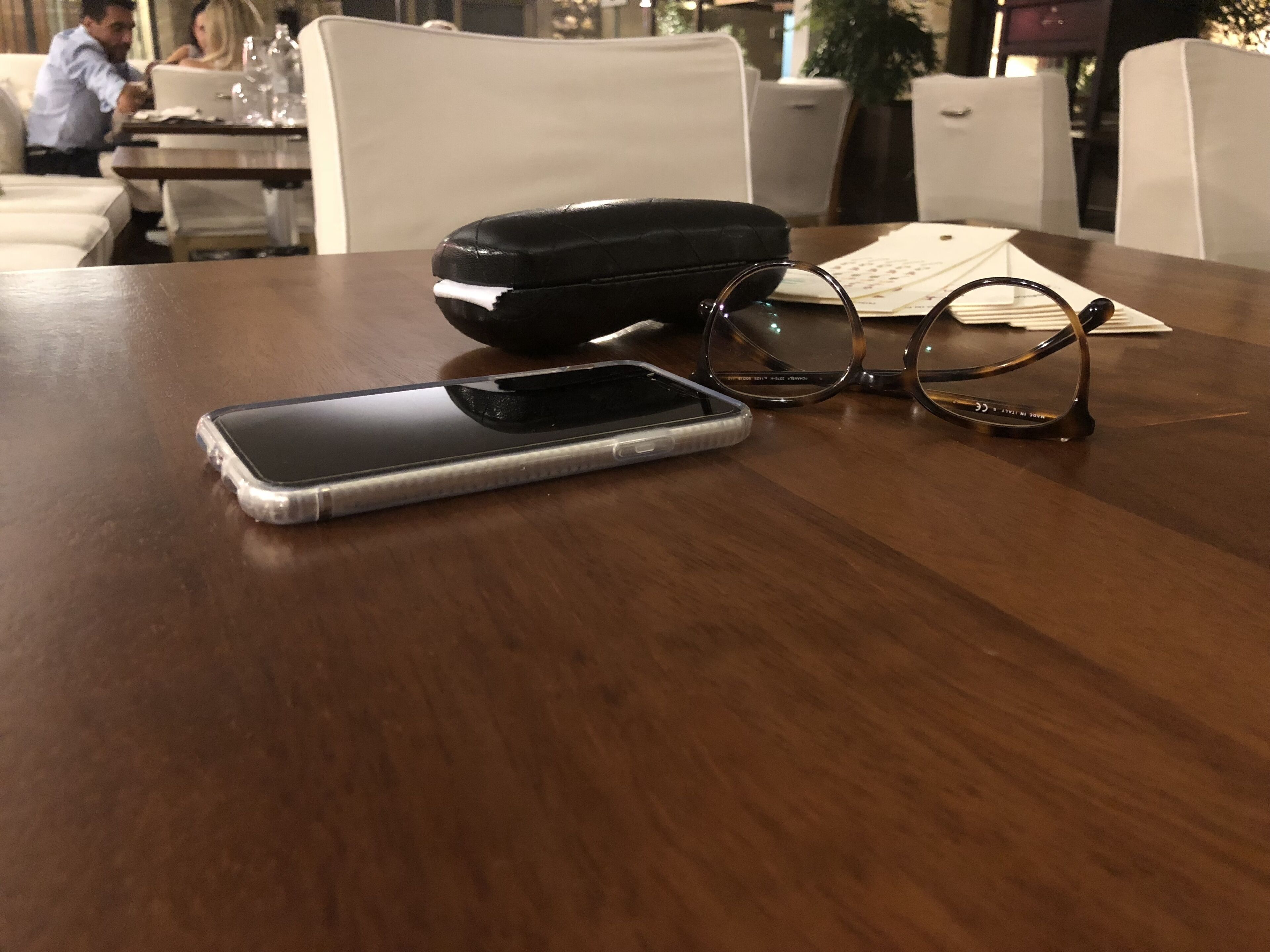 Een smartphone, een bril en een zwart hoesje op een houten tafel in een restaurant.