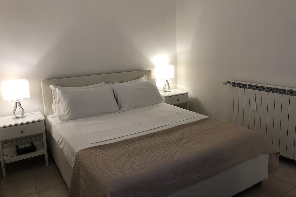 Una cama bien hecha con sábanas blancas y una manta marrón, dos mesitas de noche con lámparas y un radiador en la pared.