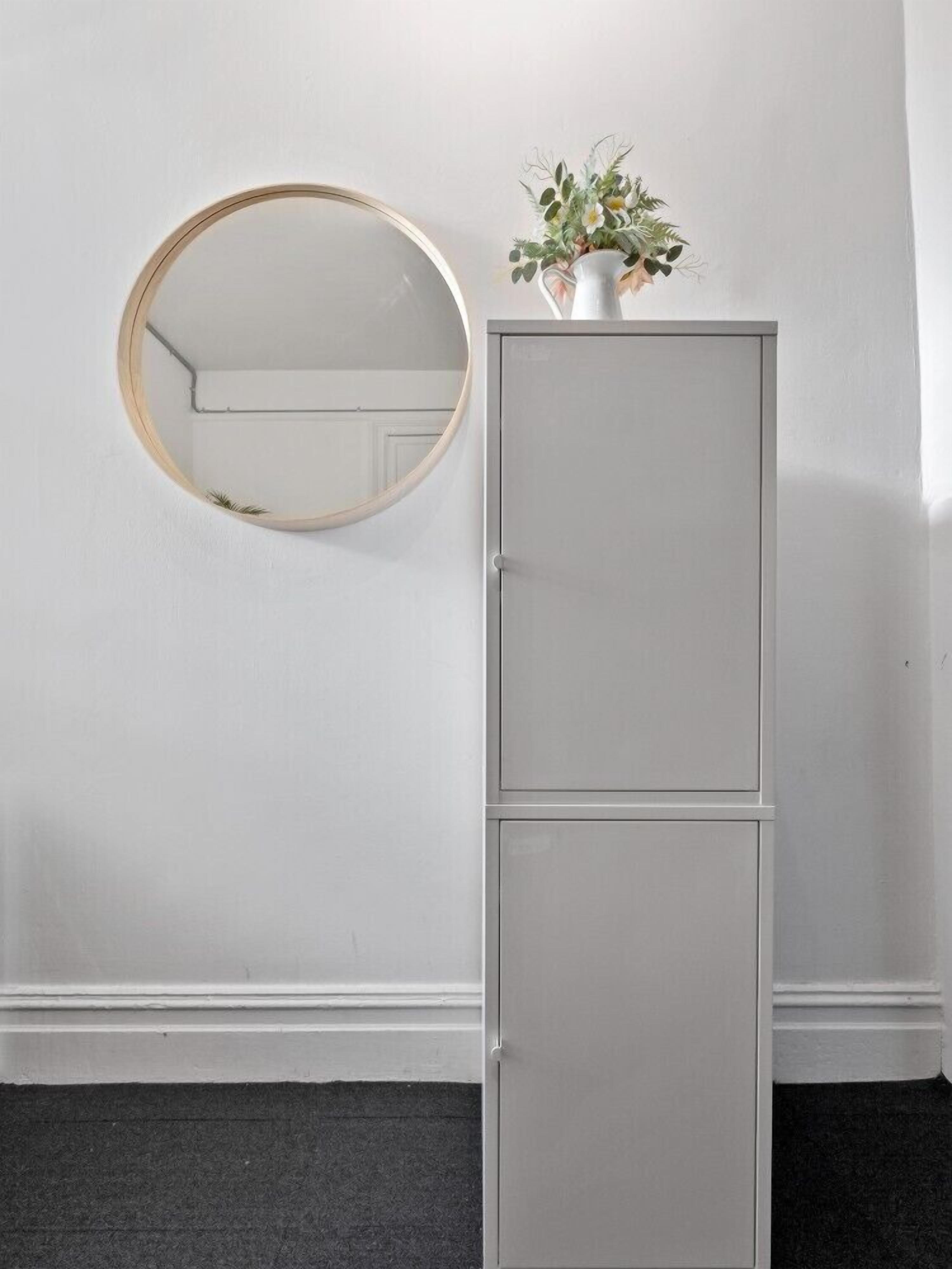 A white wall with a round mirror and a grey cabinet with a small plant on top.