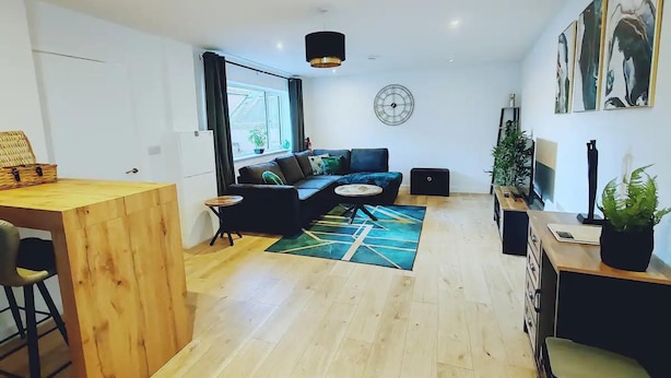A modern living room with a wooden floor, a black sofa, a small round table, and a large window. - Stunning Modern Christchurch Town Centre Apartment (Christchurch)