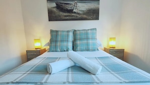 A neatly made bed with plaid bedding, two bedside tables with lamps, and a painting of a boat on the wall. - Stunning Modern Christchurch Town Centre Apartment (Christchurch)