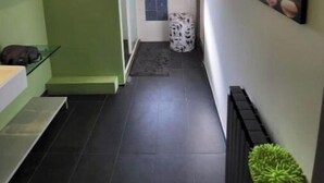 En smal gang med en grøn væg på den ene side og en hvid væg på den anden. Der er et vindue med udsigt, en skraldespand og en plante.