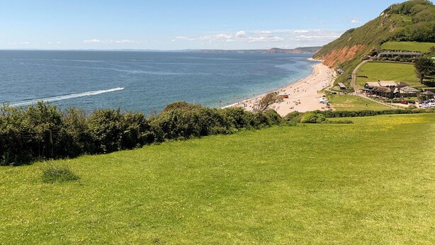 A coastal landscape with a beach, grassy fields, and a hill with buildings.