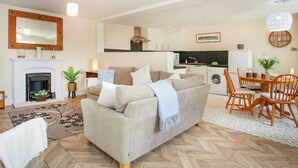 A living room with a sofa, fireplace, dining table with chairs, and a kitchen area in the background.