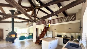 Un interior espacioso con vigas de madera a la vista, una chimenea central, una cocina con lavaplatos y una escalera que lleva a un nivel superior.