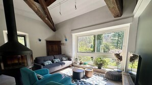 Un salón con sofá, sillas color esmeralda, chimenea, televisión y un amplio ventanal con vista a zonas verdes.