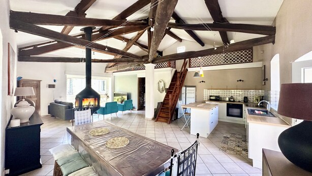 Un amplio salón con vigas de madera a la vista, una chimenea, una cocina con electrodomésticos modernos y una escalera que conduce a la planta superior.