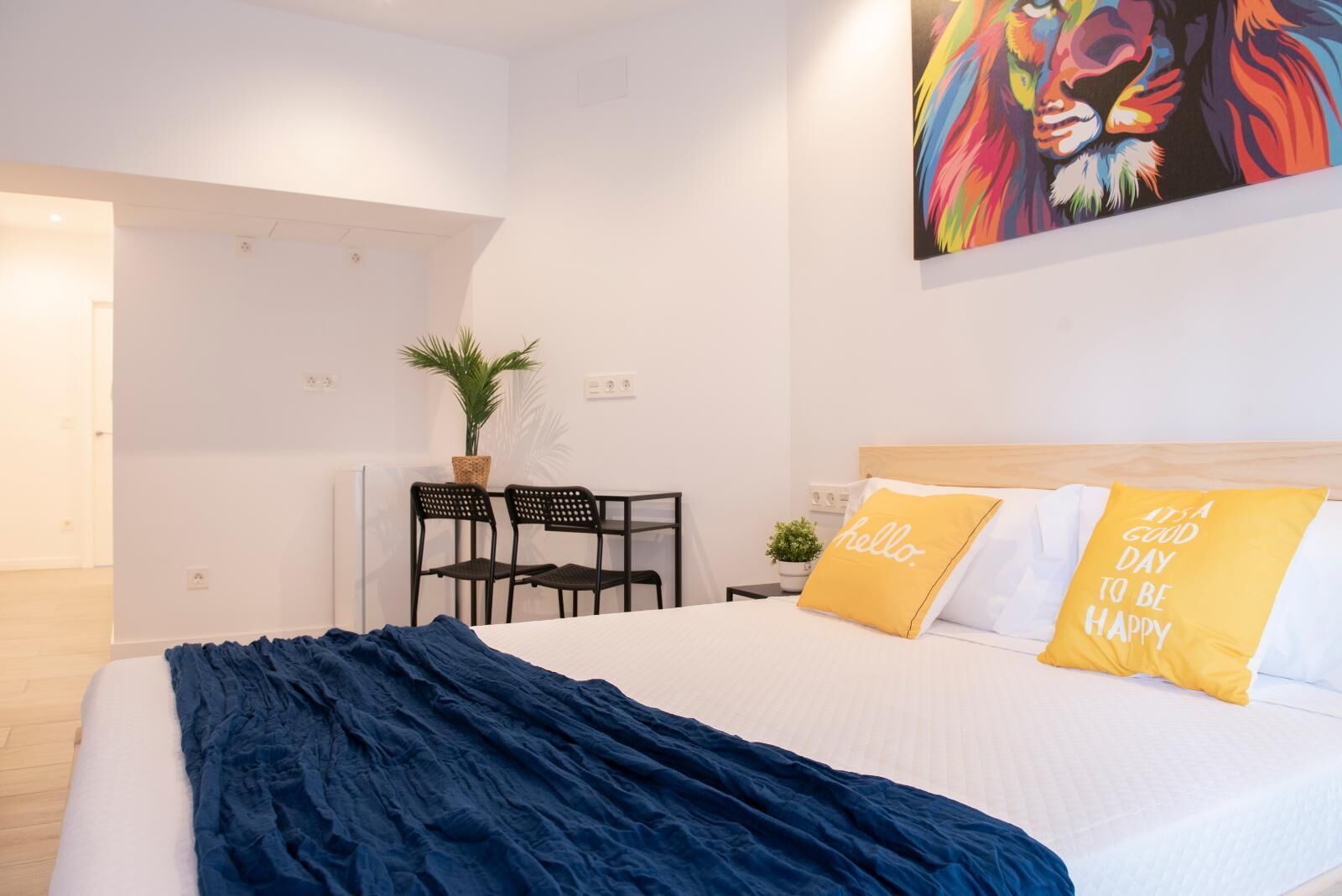 Une chambre à coucher moderne avec un lit, deux oreillers, une couverture bleue, un bureau avec des chaises et une plante.