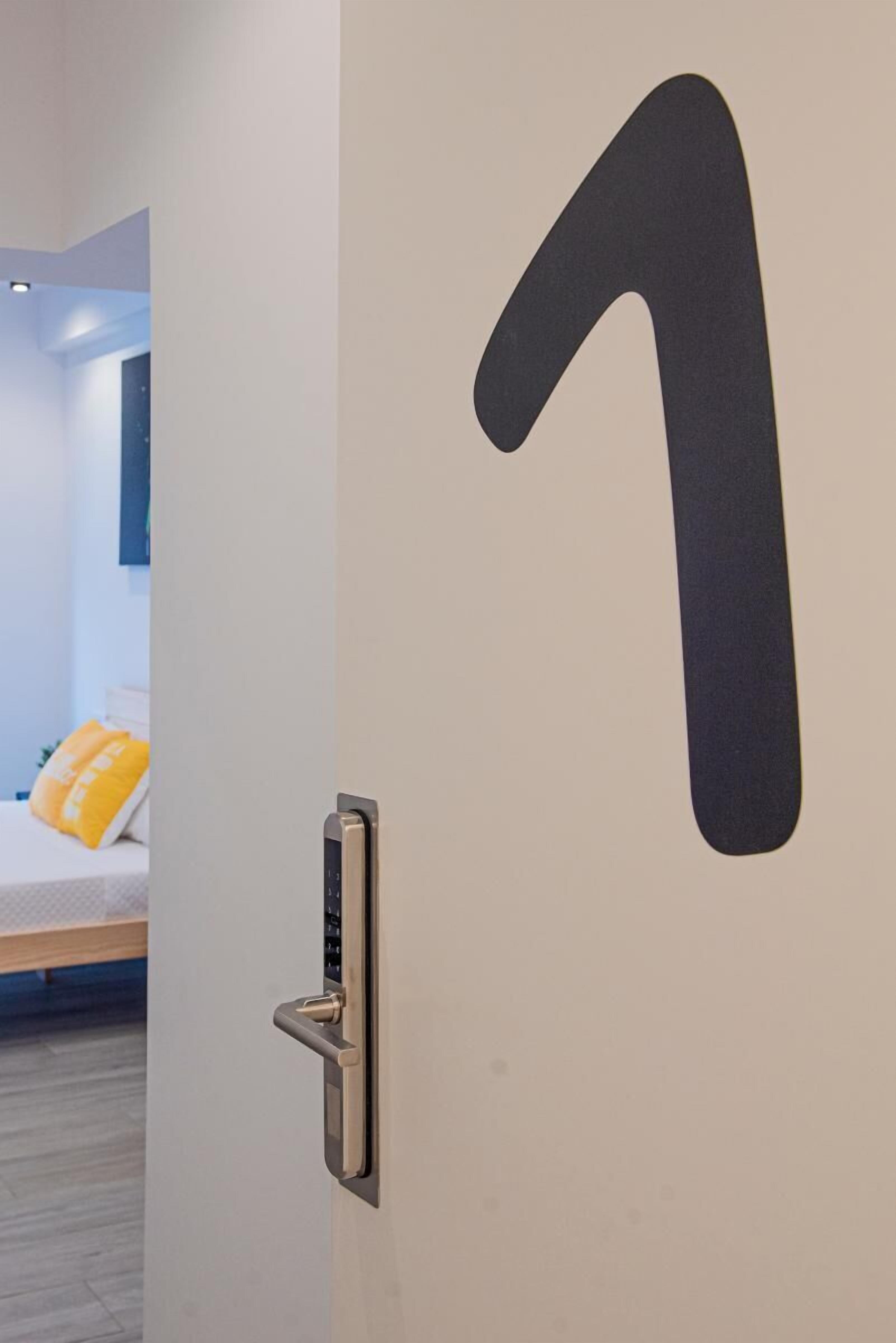 Une porte avec un grand numéro « 1 » affiché, une poignée de porte moderne, et on aperçoit une pièce avec un lit et des coussins jaunes.