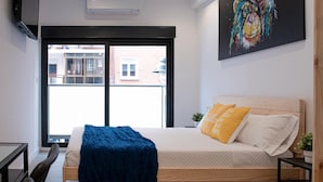A modern bedroom with a bed, a desk, and a chair. There is a large window with a view of buildings outside. - Bilbao ROOMS & KITCHEN Zorroza (Bilbao)