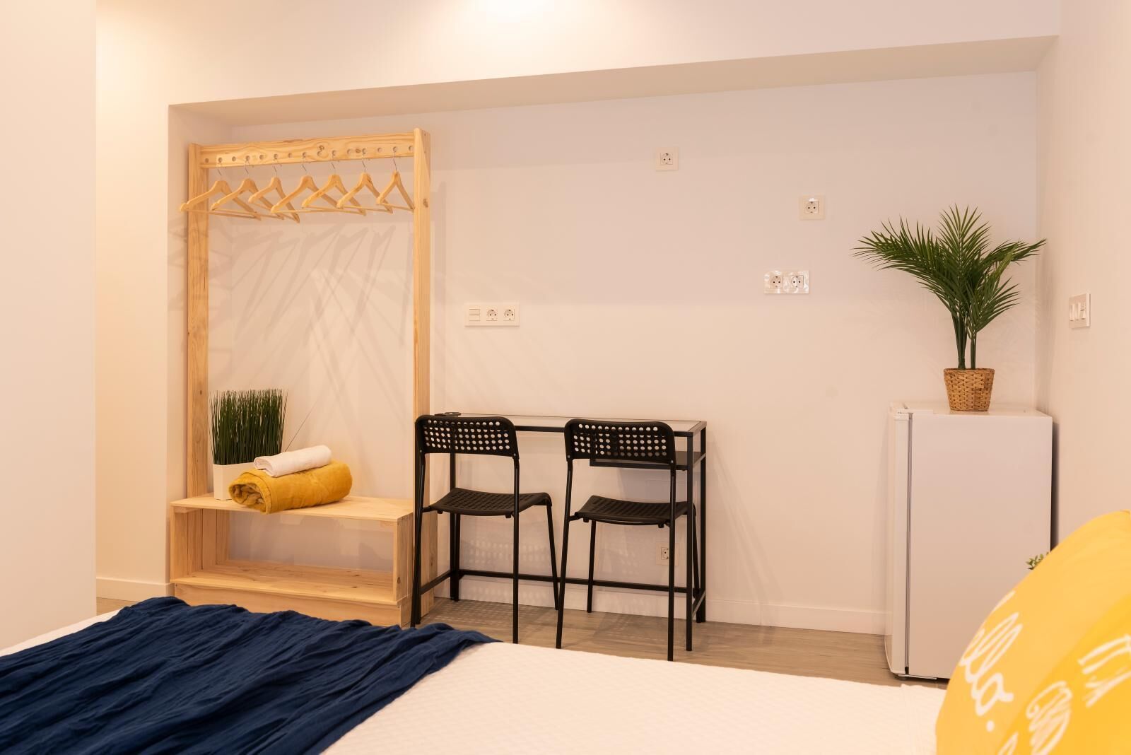 A bedroom with a bed, a desk with two chairs, a wardrobe, a small plant, and a refrigerator.