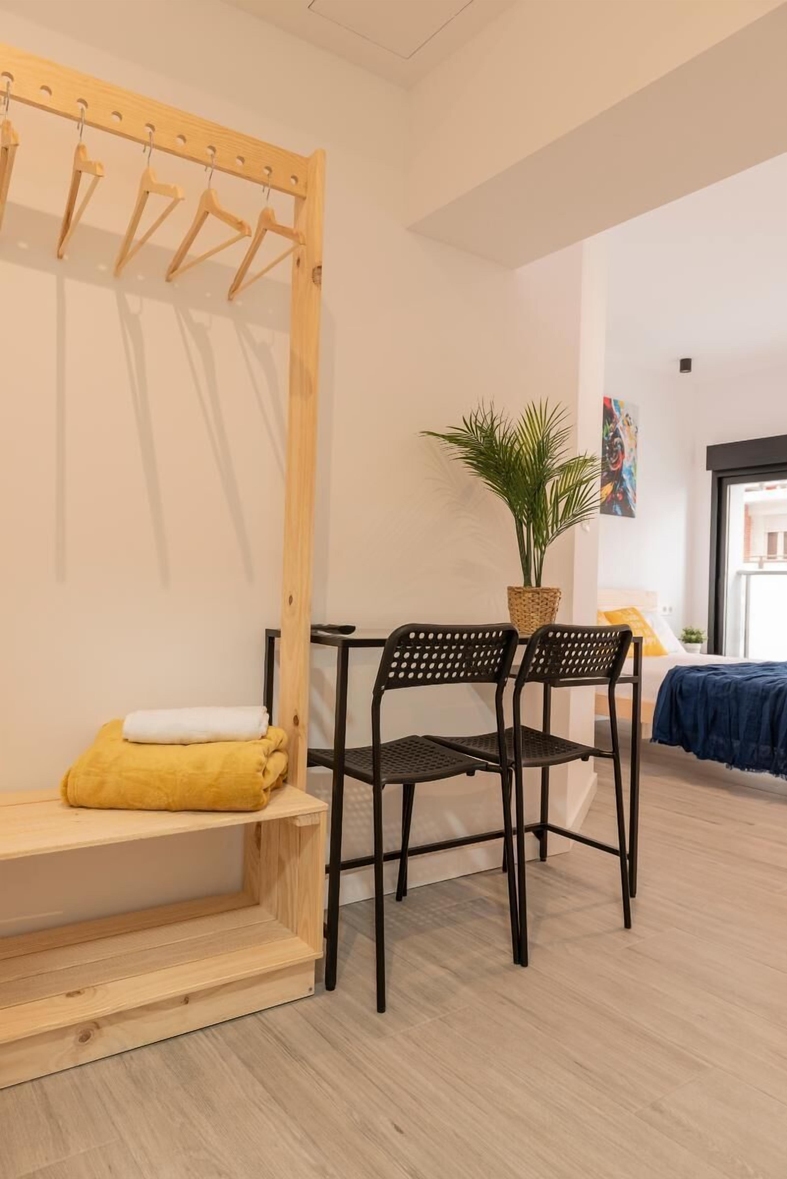 Una habitación con un banco de madera, una silla negra, una planta en maceta y una cama con una manta azul.