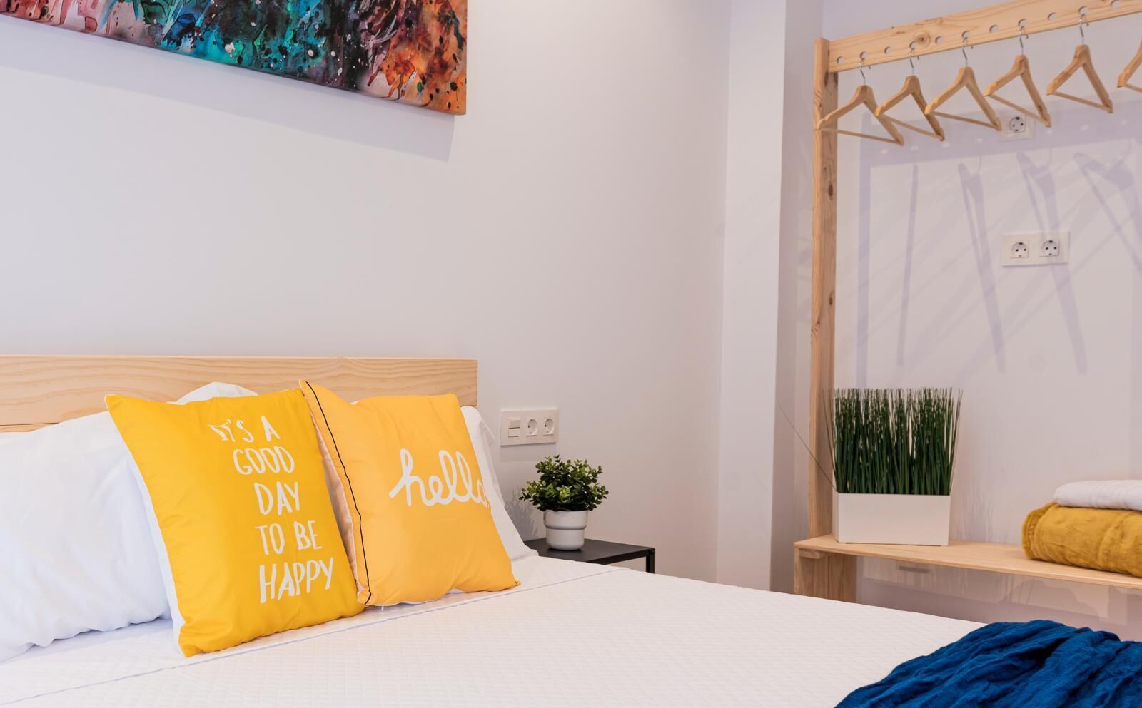 Cama bem arrumada com dois travesseiros amarelos, um travesseiro branco e um cobertor azul. Há uma pequena planta em vaso sobre uma mesinha de cabeceira e um cabideiro de madeira com roupas.