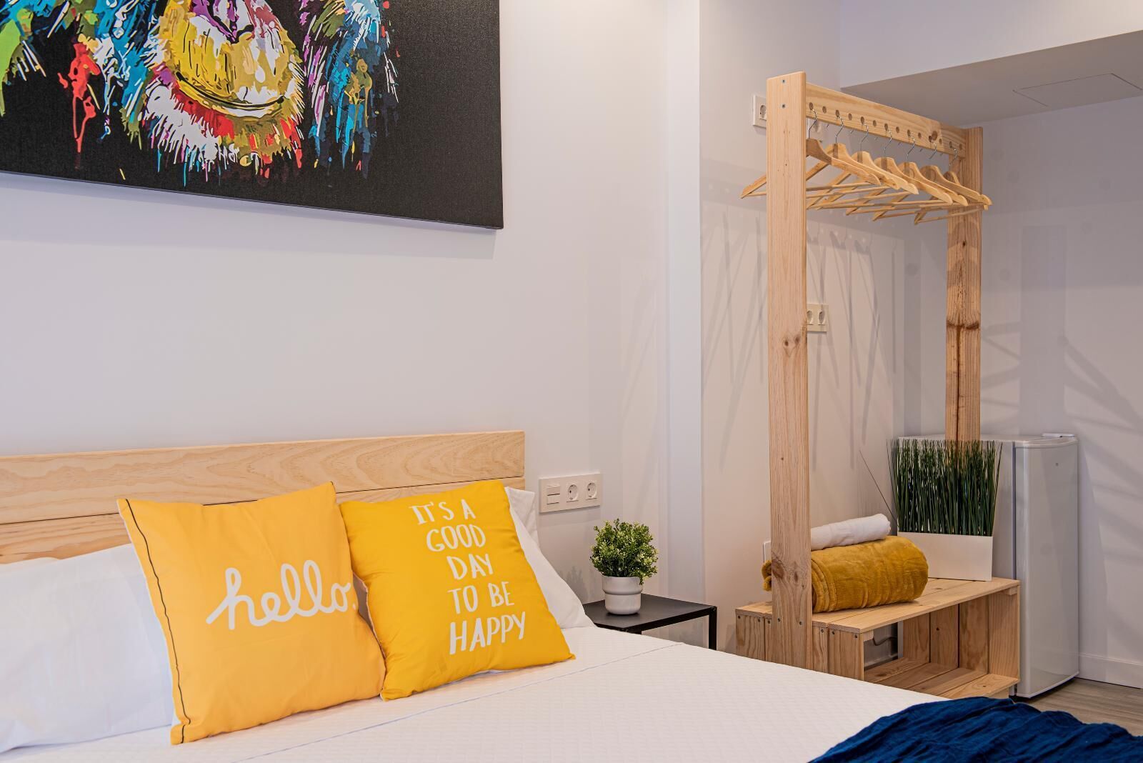 A modern bedroom with a wooden headboard, a bed with two yellow pillows, a wooden drying rack, a small potted plant, and a colorful abstract painting on the wall.