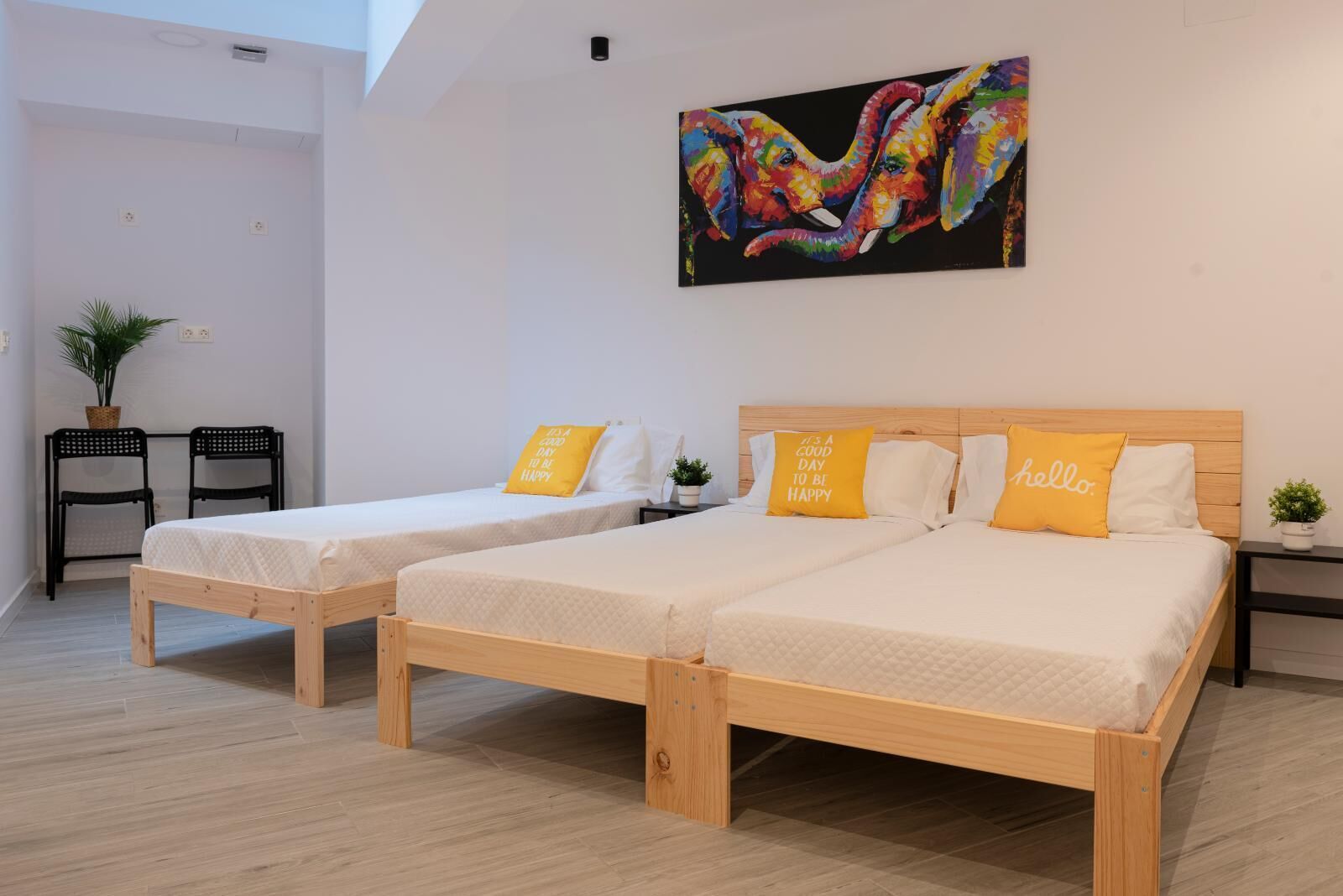 Una habitación con dos camas de madera, cada una con un colchón blanco y una almohada amarilla. Encima de las camas hay un cuadro de colores, y a la izquierda, una pequeña mesa con sillas.