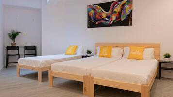 Una habitación con dos camas de madera, cada una con un colchón blanco y una almohada amarilla. Encima de las camas hay un cuadro de colores, y a la izquierda, una pequeña mesa con sillas.