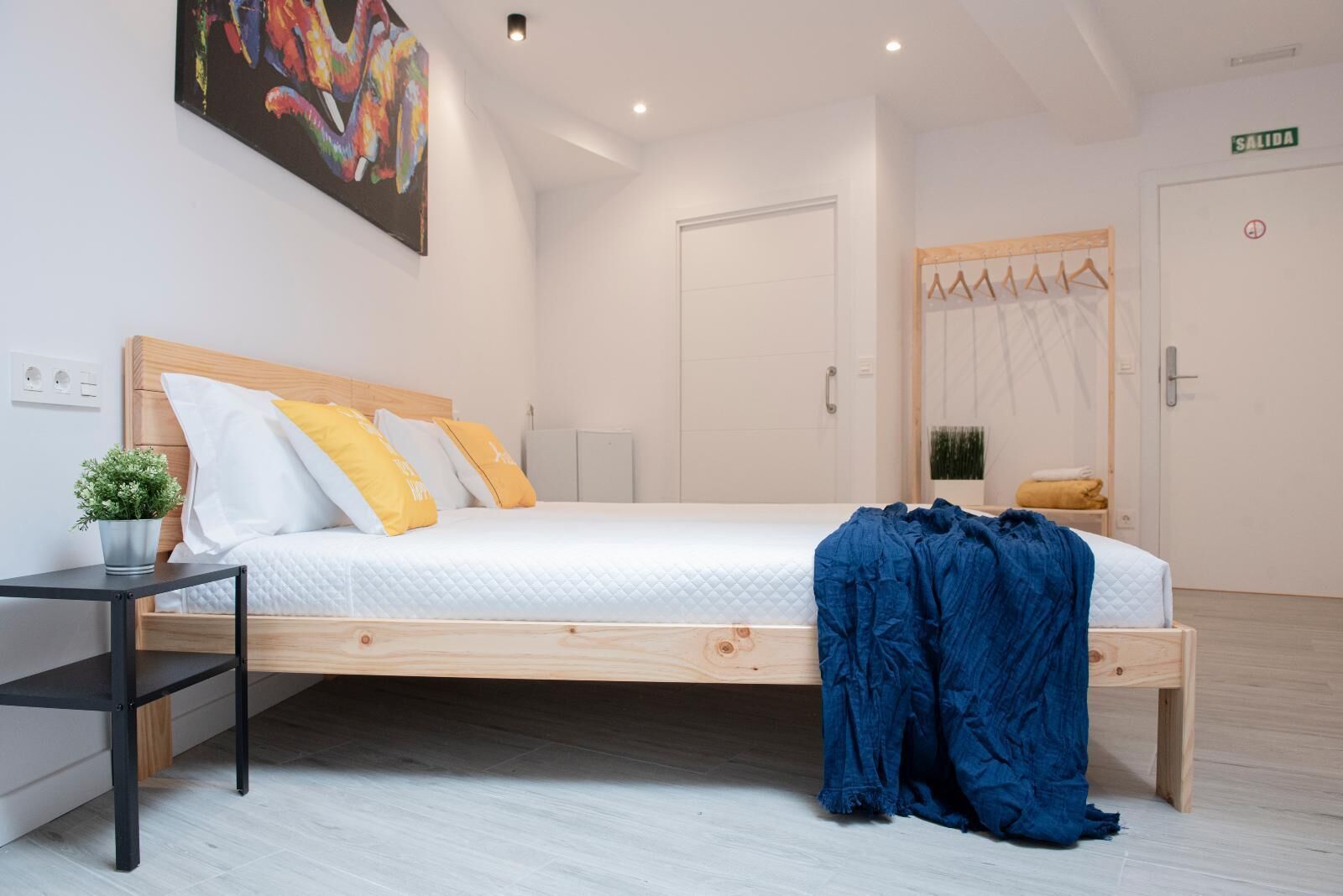 Een moderne slaapkamer met een houten bed, wit beddengoed, een klein nachtkastje met een plant, en een blauwe deken op de vloer.