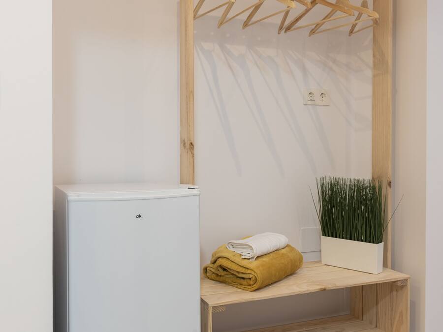 Habitación compacta con un mini refrigerador blanco, un tendedero de madera con perchas, un pequeño estante con una toalla doblada y una planta en maceta.