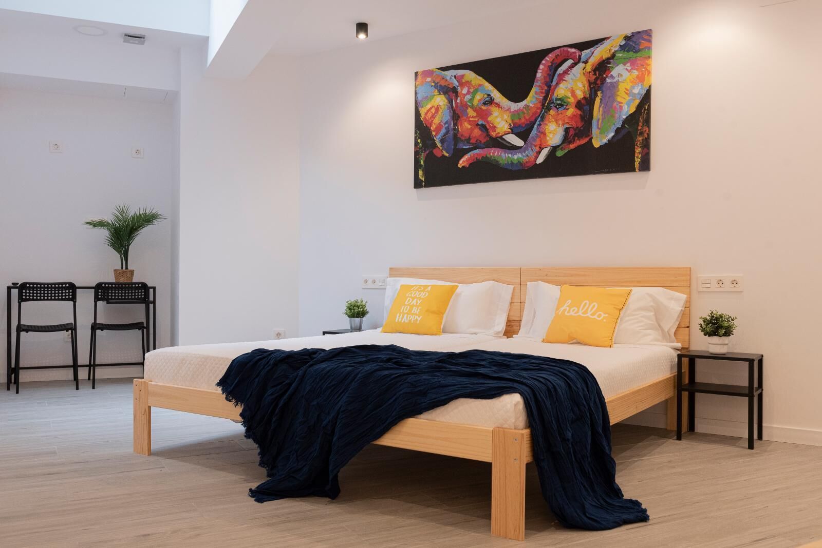 A modern bedroom with a wooden bed, two bedside tables, a wall-mounted painting, and a small potted plant.