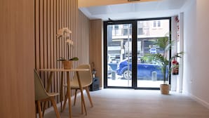 A reception area with a wooden panel wall, a reception desk with chairs, a potted plant, and a glass door leading outside. - Bilbao ROOMS & KITCHEN Zorroza (Bilbao)