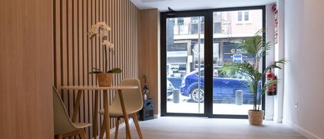 Zona de recepción con pared de paneles de madera, mostrador con sillas, planta en maceta y puerta de vidrio que da al exterior.