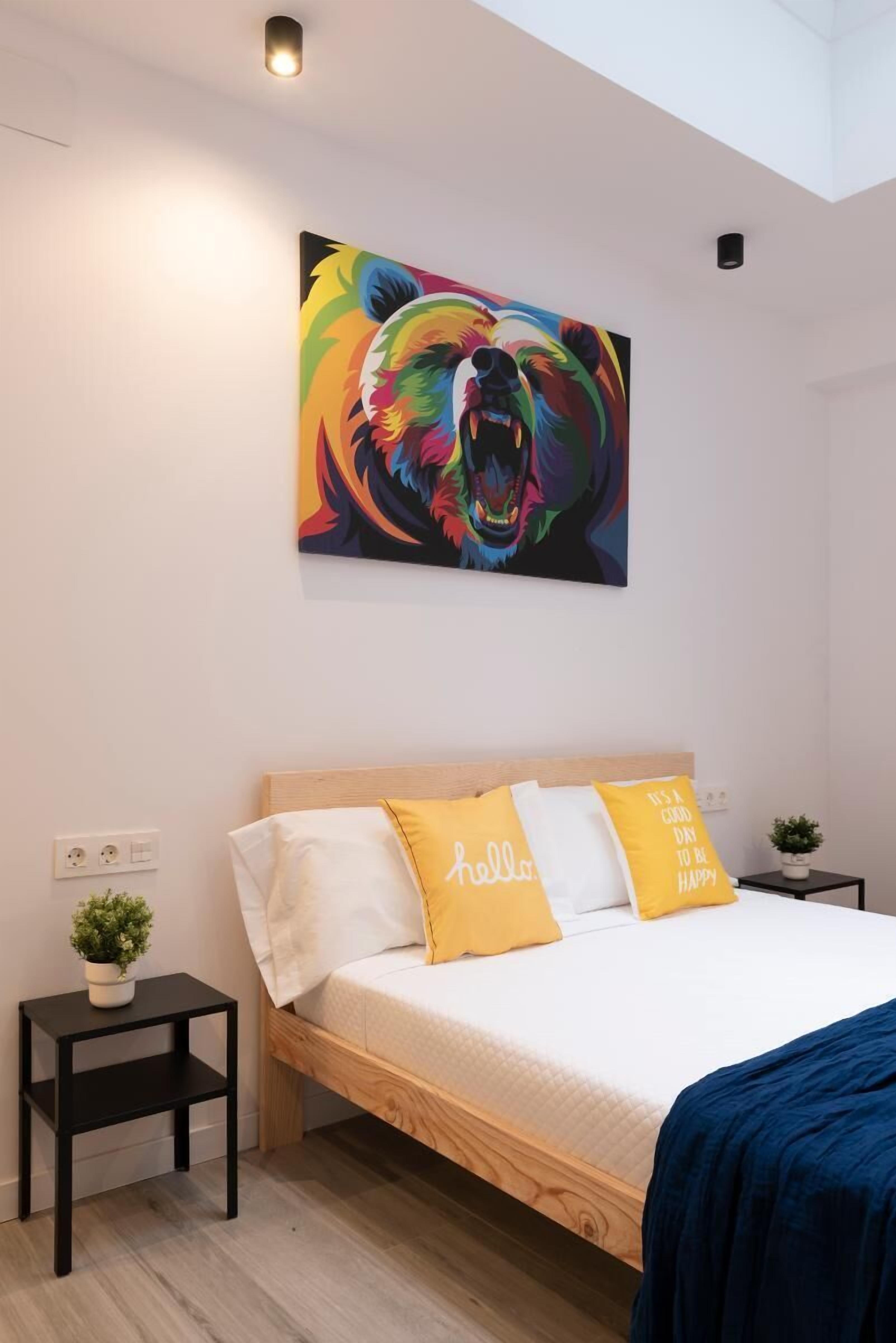 A bedroom with a wooden bed, a colorful abstract painting on the wall, and a small plant on a bedside table.