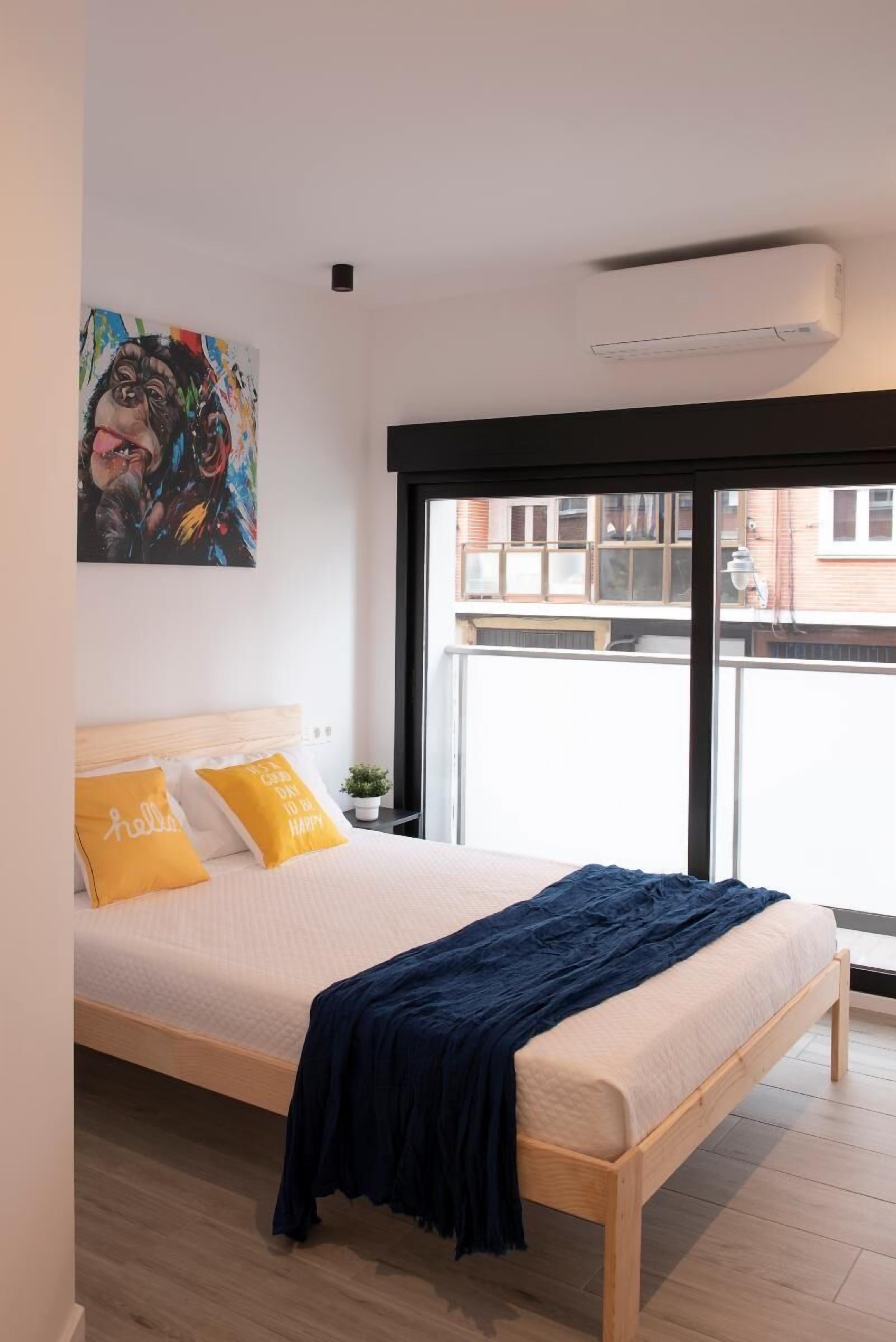 A bedroom with a large bed, a window with a view of buildings, and a colorful painting on the wall.