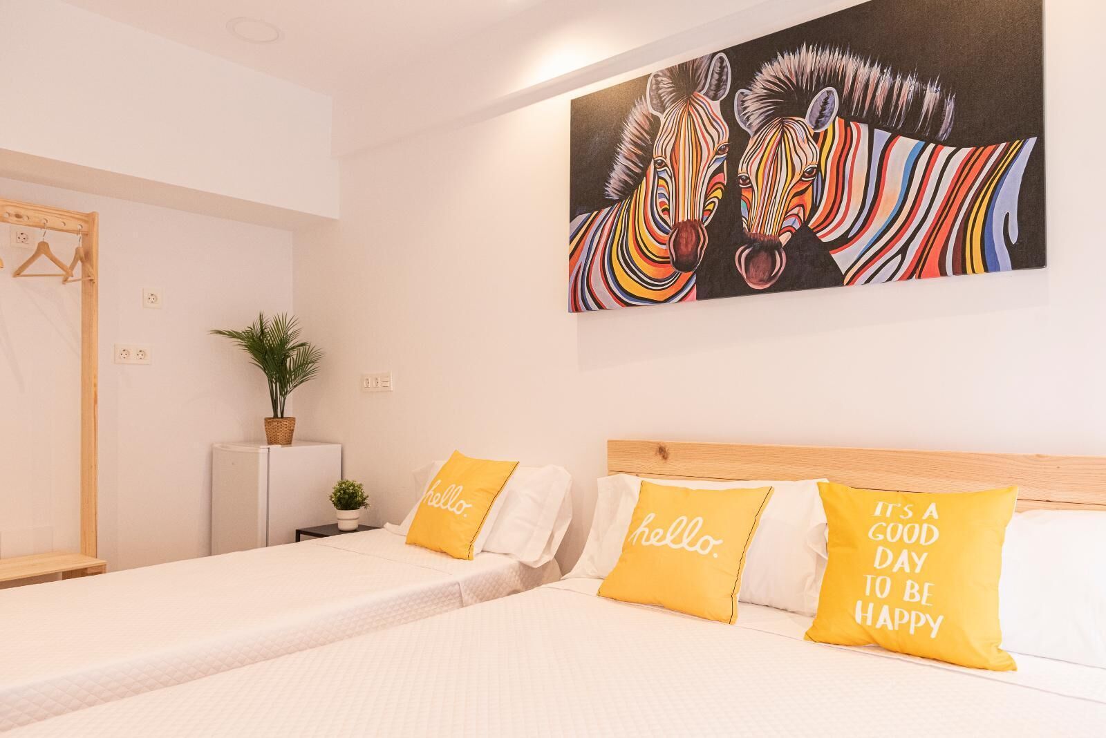 A hotel room with two beds, each with a white bedspread and a yellow pillow with "hello" and "it's a good day to be happy" text. A colorful zebra painting is on the wall above the beds.