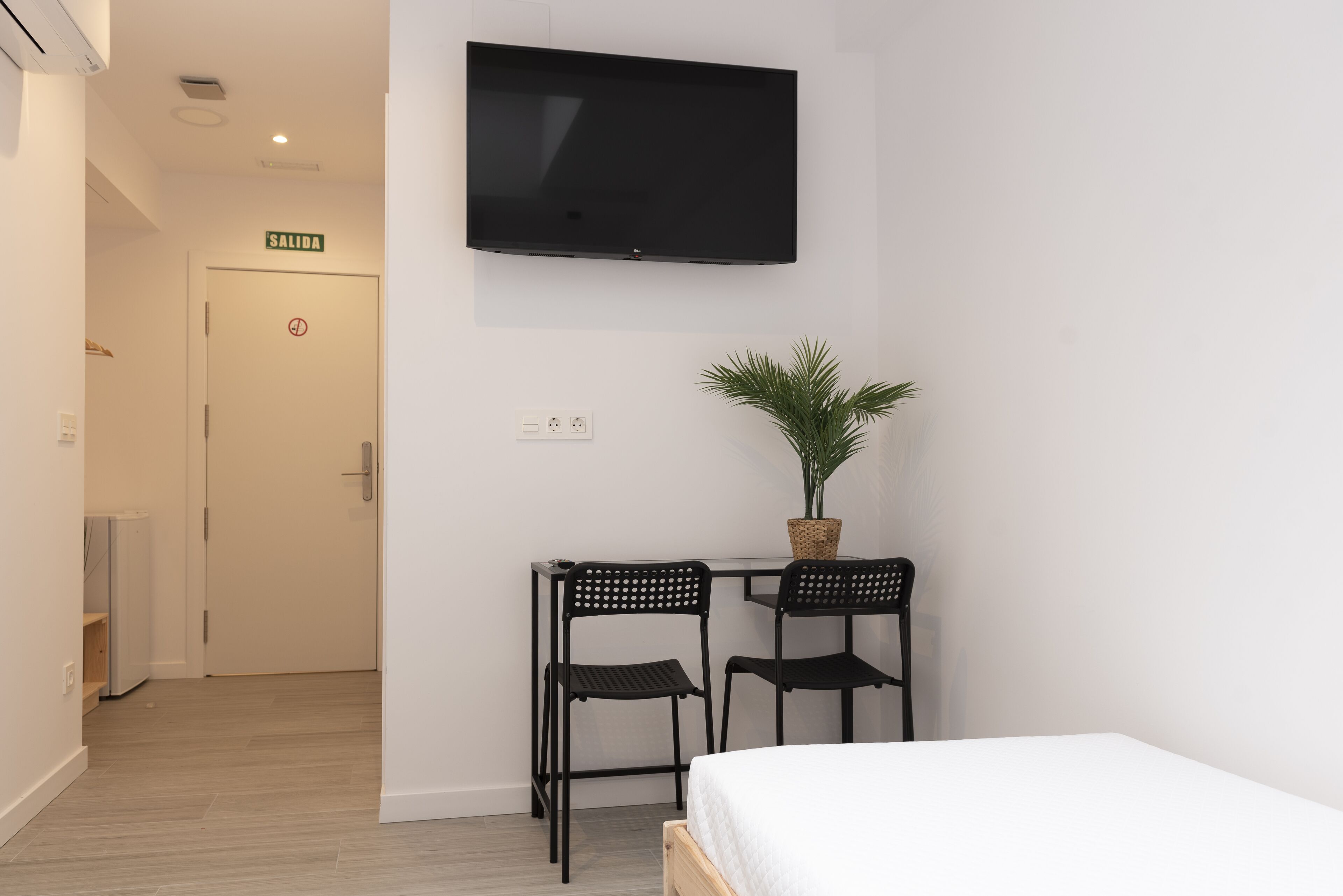 A hotel room with a bed, a TV mounted on the wall, two chairs, a small table with a plant, and a door with a 'SALIDA' sign.