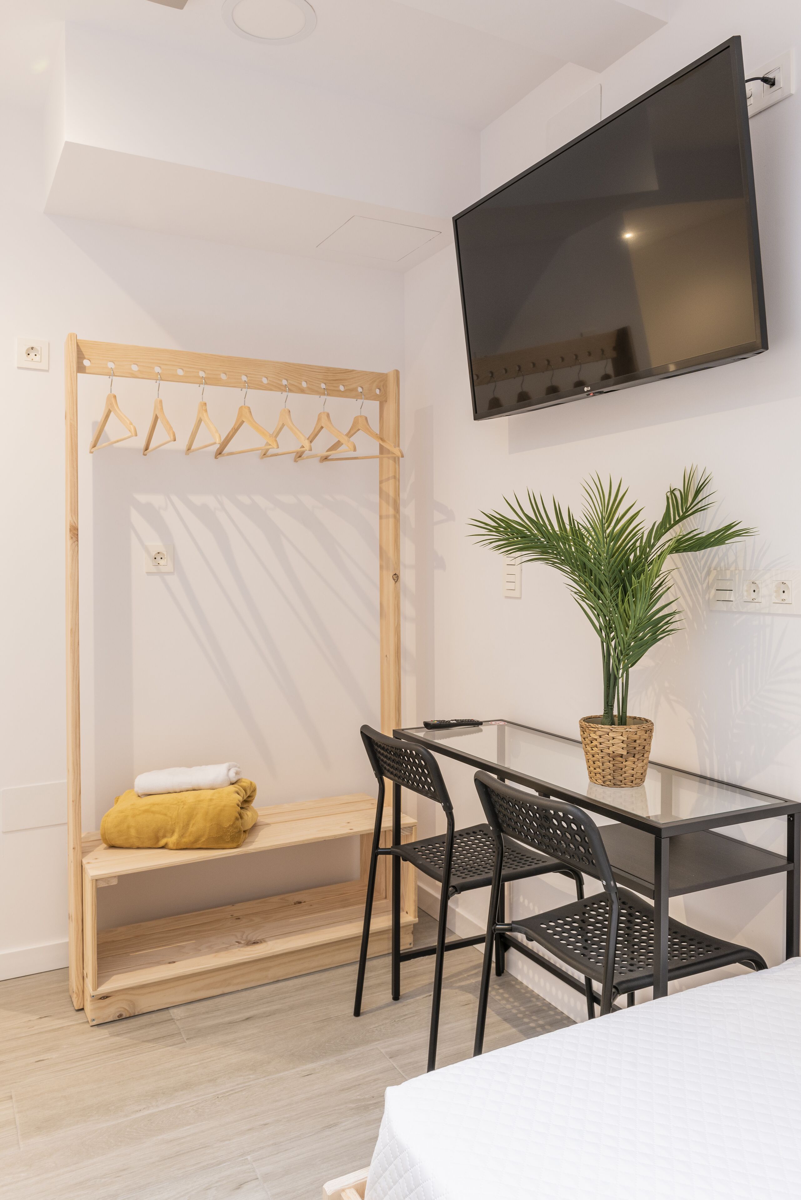 Una habitación moderna con una mesa de vidrio, dos sillas negras, un televisor montado en la pared, una silla colgante de madera con un cojín amarillo y una planta en maceta.