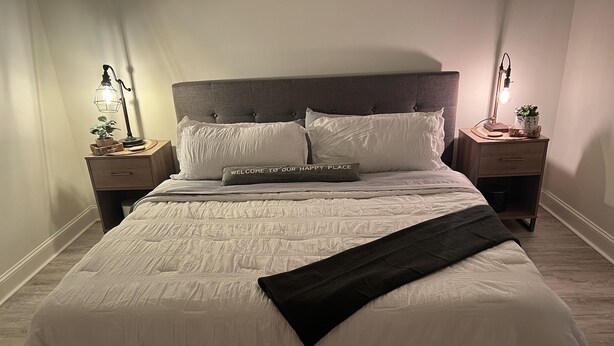 Un lit bien fait, avec une couette blanche et des oreillers, dont l’un porte l’inscription « BIENVENUE DANS NOTRE PETIT COIN DE PARADIS ». Il y a deux tables de chevet avec des lampes et une petite plante sur chacune. La pièce a un plancher en bois et une tête de lit recouverte d’un tissu gris.