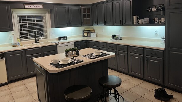 Una cocina moderna con armarios gris oscuro, una isla central con taburetes, un ventanal con persianas y una estantería con tazas y una cafetera.