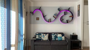 A modern living room with a black sofa, patterned cushions, a small side table, and a wall-mounted shelf with books and decorative items.