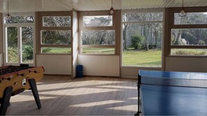 A room with a foosball table, ping pong table, and large windows offering a view of a grassy area. - Gite - 800 m from the sea & commodities in a parc of  3,5ha for familly event -  (Loctudy)
