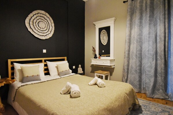 Een netjes opgemaakt bed met een beige deken, twee opgerolde handdoeken, een houten hoofdbord, een klein nachtkastje met een vaas, en een spiegel erboven.