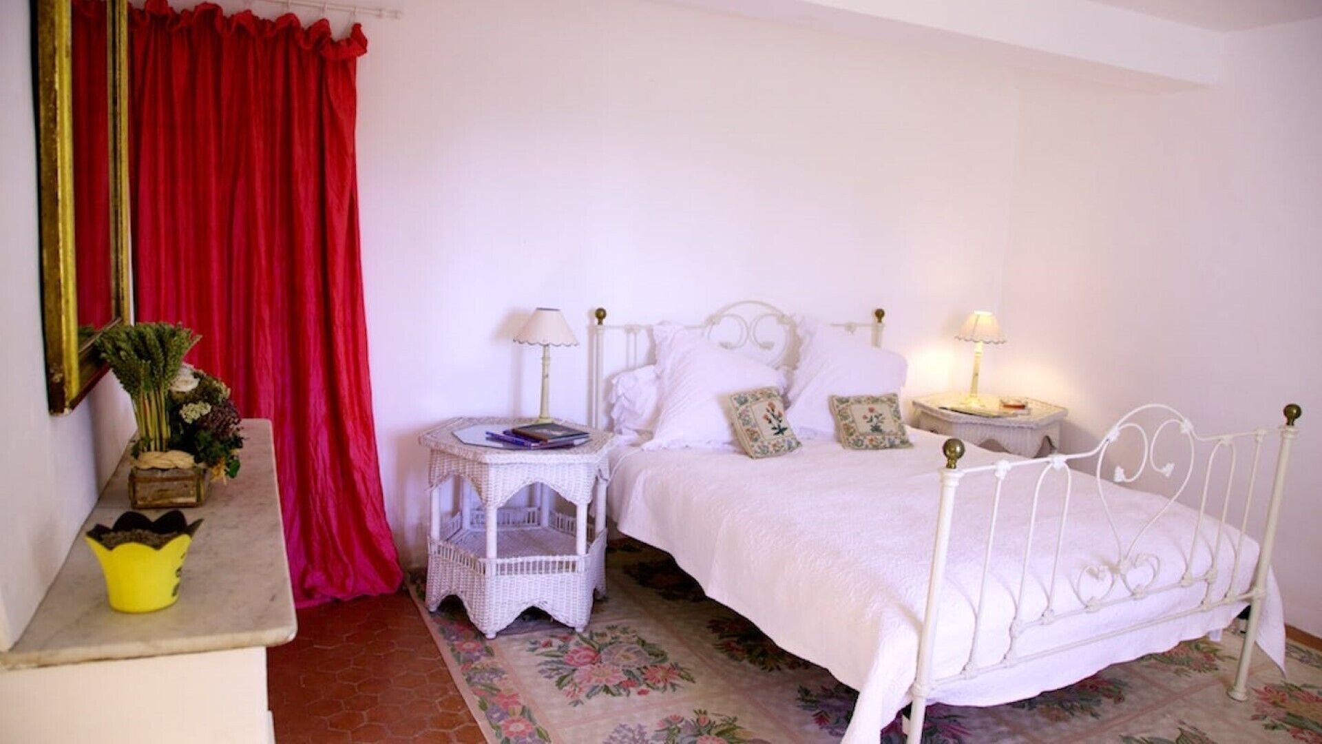 A bedroom with a white bed, two bedside tables, a red curtain, and a floral carpet.