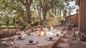 Uma área de refeições ao ar livre com uma mesa comprida posta para uma refeição, cercada por muros de pedra e árvores.