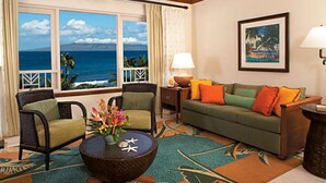 Ein Zimmer mit Meerblick, einem Deckenventilator, einem grünen Sofa mit orangen Kissen, einem Korbstuhl und einem Couchtisch mit Blumen.