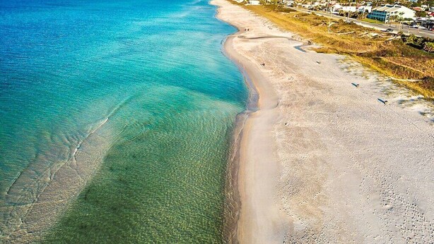 An aerial view of a beach with clear turquoise waters, a sandy shoreline, and a residential area with buildings in the background. - A Rare Nautical Historic Gem! Steps to the Ocean! Grocery Next Door! (Panama City Beach)