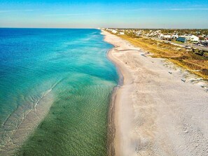 An aerial view of a beach with clear turquoise waters, a sandy shoreline, and a residential area with buildings in the background.