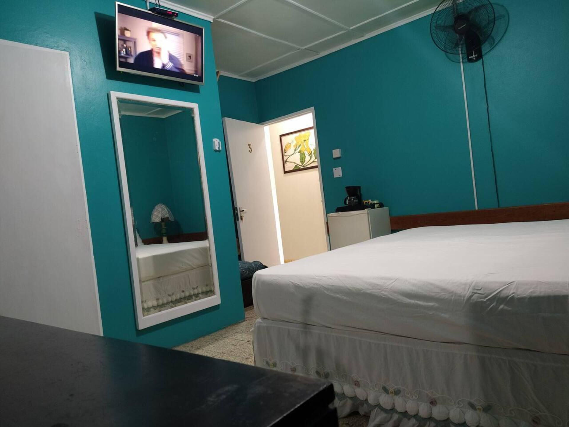 A room with teal walls, a bed, a TV on the wall, a fan, and a small kitchenette.